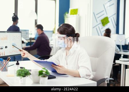 Manager wearing protection against covid 19 during global pandemic. Employees with visors working in corporate company workspace respecting social distance analysing data and graphs. Stock Photo