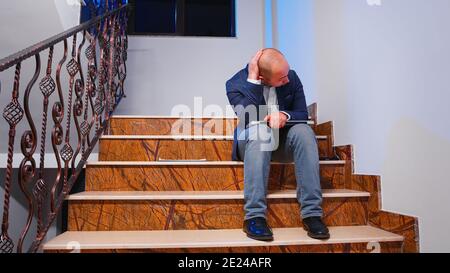 Company manager working late at night finishing business deadline project closing laptop and checking documents from clipboard sitting on stairs from corporate building. Stock Photo