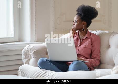 Afro American millennial woman with afro hairstyle sitting on couch taking break from work on laptop Stock Photo