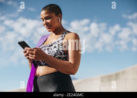 Female in sportswear using her smart phone after workout session. Healthy woman wearing wireless earphones taking a break from workout and checking he Stock Photo