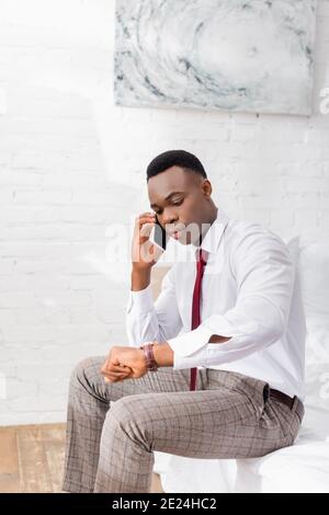 African american businessman checking time while talking on smartphone on bed Stock Photo
