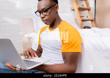 African american freelancer in eyeglasses using laptop on blurred foreground in bedroom Stock Photo