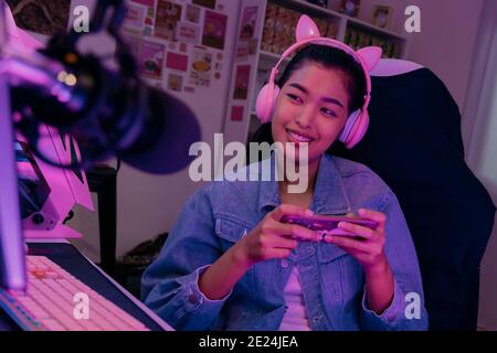 Young playful Asian woman sitting in front of her computer workstation and smiling at the screen with a microphone in foreground. Beautiful asian woman working from her home studio and playing game. Stock Photo