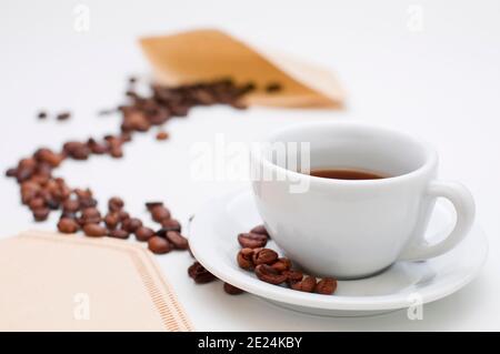 White half-filled cappuccino cup with saucer and individual roasted coffee beans in the sharp foreground and coffee beans arranged in an arch towards