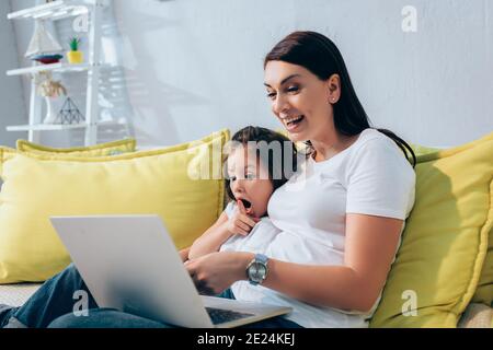 Excited daughter and mother laughing while looking at laptop on couch on blurred background Stock Photo