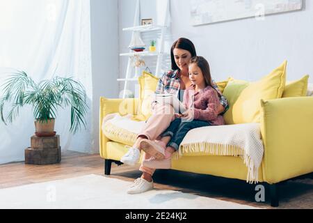Full length of happy mother and daughter looking at copy book while sitting on couch in living room Stock Photo