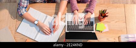 Cropped view of daughter typing on laptop near mother at desk with stationery, banner Stock Photo