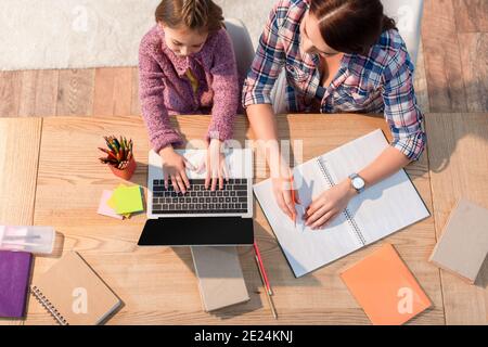 Top view of mother sitting near daughter typing on laptop at desk with stationery at home Stock Photo