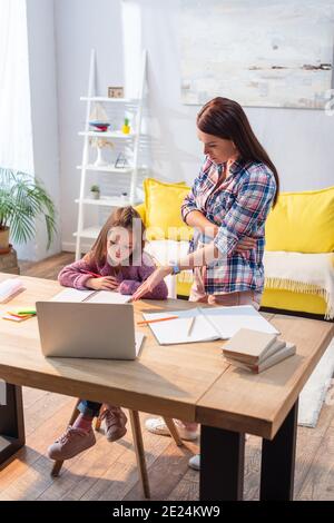 Full length of serious mother pointing with hand at laptop near upset daughter at desk in living room Stock Photo