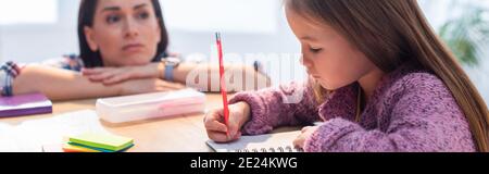 Worried mother looking at daughter writing in notebook at desk on blurred background, banner Stock Photo
