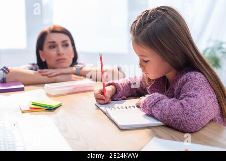 Upset mother looking at daughter writing in notebook at desk on blurred background Stock Photo