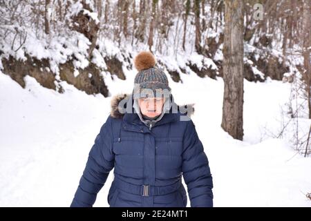 Retired woman in jacket and hat with bright colors looking where she steps on a snow-filled path in the forest. Scene after the snowstorm called Filom Stock Photo