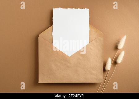 Envelope with blank paper card inside and dried flowers on brown background. Flat lay, top view. Stock Photo