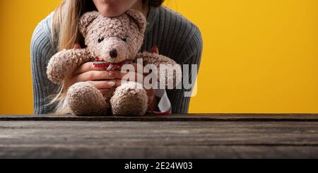 Young woman leaning her head on a vintage teddy bear toy. Over yellow background with copy space. Stock Photo