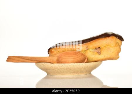 One chocolate eclair on a ceramic saucer with a wooden spoon, close-up, isolated on white. Stock Photo