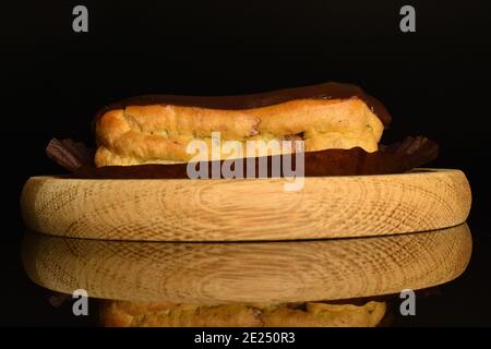 One chocolate eclair on a wooden tray, close-up, on a black background. Stock Photo