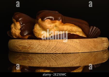 Two chocolate eclairs on a wooden tray, close-up, on a black background. Stock Photo