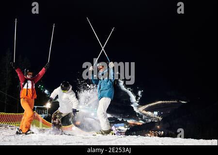 Friends skiers in winter jackets and helmets having fun at ski resort in the mountains in winter, skiing at night, illuminated ski pistes are downhill on background. Concept of healthy lifestyle Stock Photo