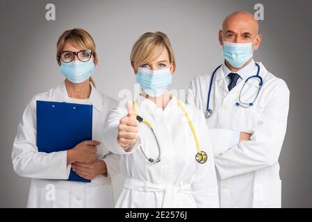 Shot of female and male doctors standing together at isolated grey background while female doctor giving thumbs up. Medical team. Doctors wearing face Stock Photo