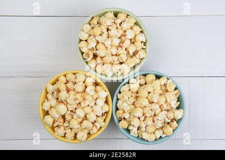 Overhead shot of three different colored bowls filled with fresh popped popcorn, on a white wood table. Stock Photo