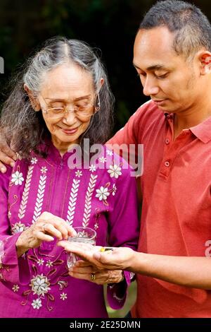 Southeast Asian ethnic senior woman taking pills helped by her young adult son Stock Photo