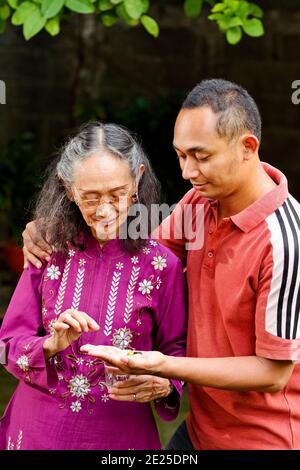 Southeast Asian ethnic senior woman taking pills helped by her young adult son Stock Photo