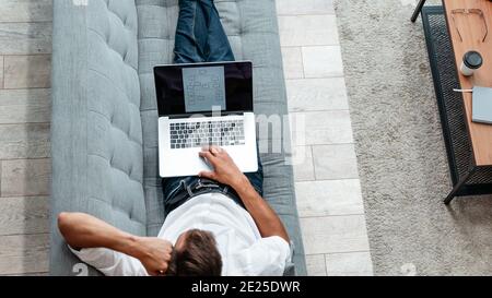 male freelancer is working on a laptop while lying on the couch . Stock Photo