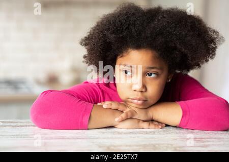 Portrait of cute sad little black girl sitting at table in kitchen. Stock Photo