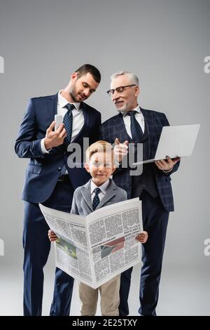 mature businessman talking to son and pointing with finger at grandson holding newspaper on grey Stock Photo