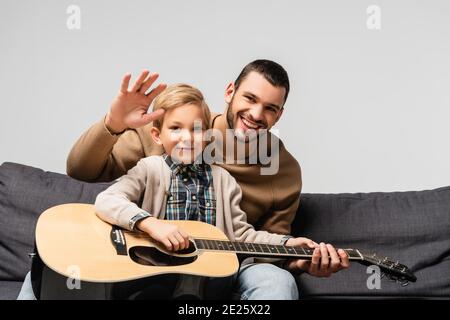 cheerful boy playing acoustic guitar near happy father waving hand at camera isolated on grey Stock Photo