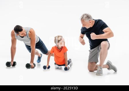 happy grandfather demonstrating strength near father and kid doing push ups with dumbbells on white Stock Photo