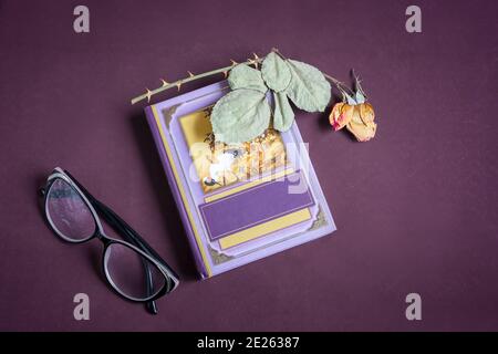 On a small book is a dried rose flower with leaves. There are glasses next to it. Presented on a dark background. Top view with space to copy, flat la Stock Photo
