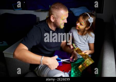 Daughter and father having fun making craft together at home on the sofa, cutting a paper with scissors, dark light, Fathers day, family. Stock Photo