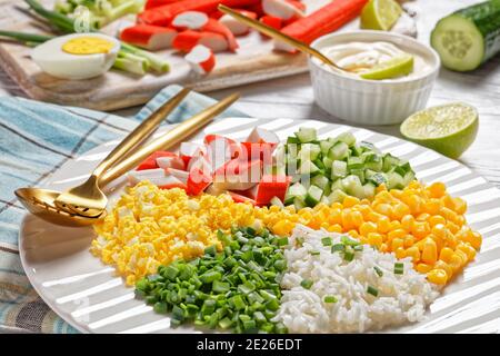 Crab salad of surimi sticks with canned corn, cucumbers, spring onion hard-boiled eggs, jasmine rice divided into sectors on a white plate on a white Stock Photo