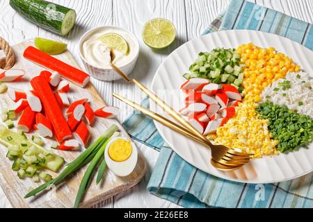 Crab salad of surimi sticks with canned corn, cucumbers, spring onion hard-boiled eggs, jasmine rice divided into sectors on a white plate on a white Stock Photo