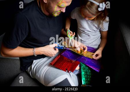 Daughter and father having fun making craft together at home on the sofa, cutting a paper with scissors, dark light, Fathers day, family. Stock Photo