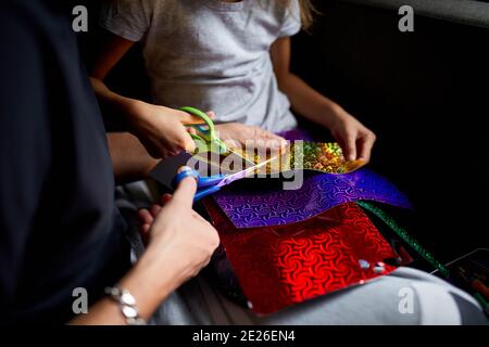 Daughter and father having fun making craft together at home on the sofa, cutting a paper with scissors, dark light, Fathers day, family. Stock Photo