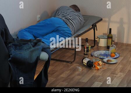 A drunk man lies on a cot and an empty bottle of alcohol is scattered nearby. Stock Photo