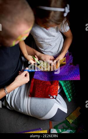 Daughter and father having fun making craft together at home on the sofa, cutting a paper with scissors, dark light, Fathers day, family. Stock Photo