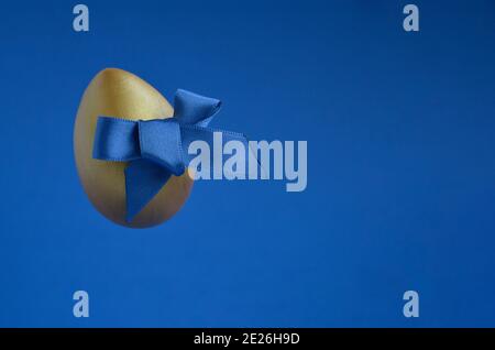 Easter golden egg with a blue bow on a blue background with space for text, levitation. Banner, poster. Stock Photo