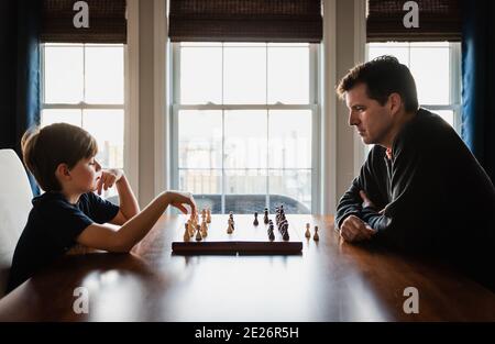 Father and son sitting at a table indoors playing a game of chess. Stock Photo