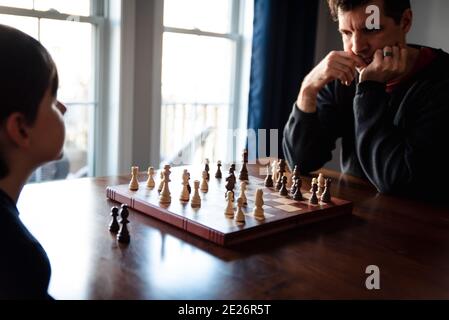Father and son sitting at a table indoors playing a game of chess. Stock Photo