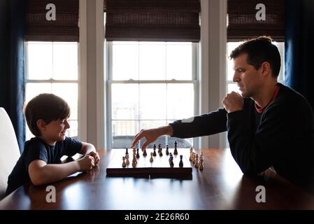 Father and son sitting at a table indoors playing a game of chess. Stock Photo