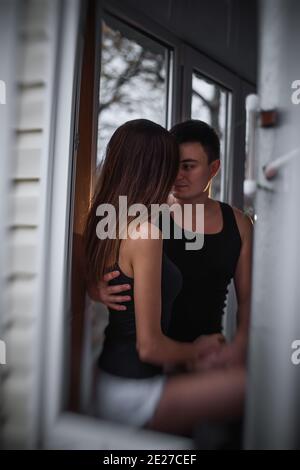 A loving young couple hugs in the window of their balcony terrace in the apartment. Young man laughs with a beautiful woman at home. Lovers kiss, pass Stock Photo