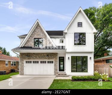 A new, white modern farmhouse with a dark shingled roof and black windows. The left side of the house is covered in a rock siding. Stock Photo