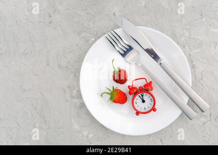 Red ripe strawberries berry on white plate, cutlery and red alarm clock on gray stone background table. Top view, flat lay, copy space Template. Conce Stock Photo