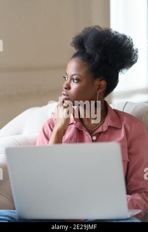 Afro American millennial woman with afro hairstyle sitting on couch taking break from work on laptop Stock Photo