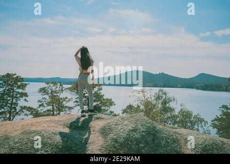 Young beautiful girl in sportswear stands on the top of mountain and looks at the lake and mountains against the blue sky. Outdoor activity or hiking Stock Photo