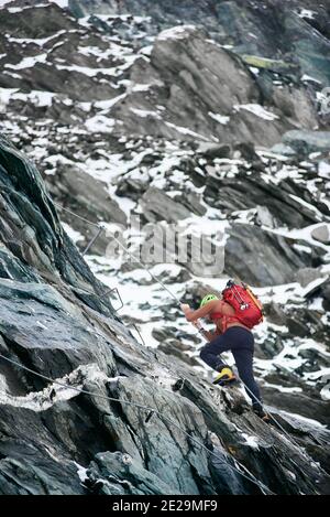 Male alpinist with backpack using rope while climbing alpine ridge. Young traveler ascending mountain cliff and trying to reach mountaintop. Concept of mountaineering, alpinism and alpine climbing. Stock Photo