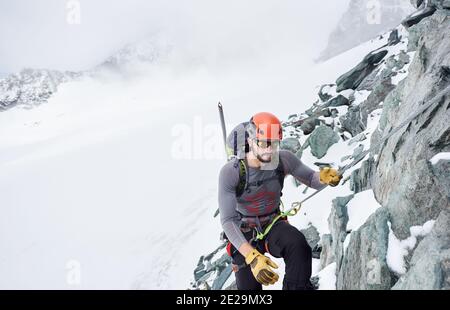 Man alpinist in sunglasses and safety helmet holding fixed rope while climbing snowy mountain. Mountaineer ascending natural rock formation. Concept of mountaineering and winter rock climbing. Stock Photo